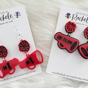 May include: Two pairs of red and black acrylic earrings. One pair features a red megaphone with the text "#1" and a red and black pom-pom. The other pair features a red megaphone with the text "GO TEAM" and a red and black pom-pom.