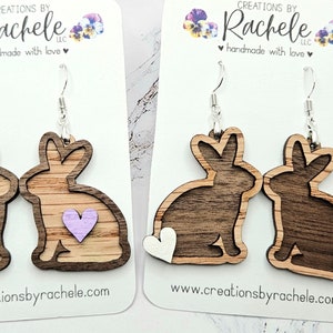 May include: Two pairs of wooden rabbit earrings on display cards. The first pair features light wood bunnies with small lavender hearts. The second pair features dark wood bunnies with white heart-shaped tails. Each pair has silver-toned metal hooks.
