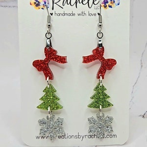 May include: A pair of silver dangle earrings with a red glitter bow, green glitter Christmas tree, and silver glitter snowflake charm. The earrings are handmade with love.