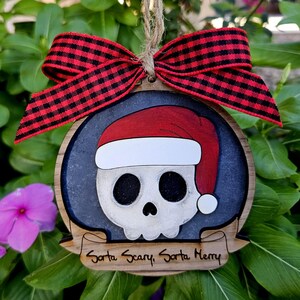 Puede incluir: Adorno navideño de madera con una calavera con un gorro de Papá Noel. La calavera es blanca con cuencas de ojos y nariz negras. El adorno tiene un lazo a cuadros rojo y negro y el texto "Santa Scary, Santa Merry".