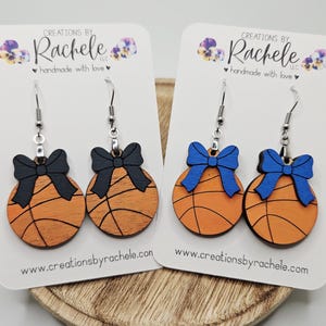 Puede incluir: Dos pares de pendientes con temática de baloncesto. Cada pendiente es un diseño de baloncesto de madera con detalles de lazo negro o azul. Los pendientes se muestran en tarjetas blancas con el logotipo y el sitio web del fabricante.