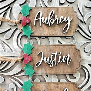 May include: Three wooden Christmas ornaments with holly and berries. The ornaments are personalized with the names Aubrey, Justin, and Jacob.
