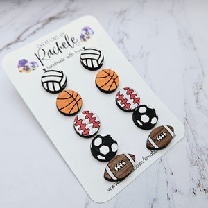May include: A set of eight wooden earrings featuring different sports ball designs. The earrings are painted in various colors, including black, white, red, orange, and brown. The earrings are arranged on a white card with the text "Creations by Rachele handmade with love" printed on it.