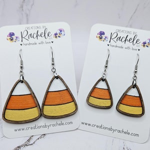 Puede incluir: Un par de pendientes de madera con forma de maíz dulce, pintados en naranja, amarillo y blanco. Los pendientes están en una tarjeta blanca con el texto "Creations by Rachele LLC handmade with love" y el sitio web "www.creationsbyrachele.com".