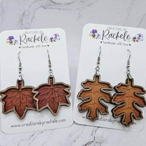 May include: Two pairs of wooden leaf earrings with silver-colored hooks. One pair features a maple leaf design in shades of brown, and the other has an oak leaf design in shades of orange and brown. The earrings are displayed on white cards with the text "Creations by Rachele".
