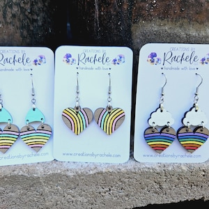 May include: Three pairs of wooden earrings with rainbow heart designs. The first pair has a light blue top piece, the second pair has a rainbow heart, and the third pair has a white cloud top piece.