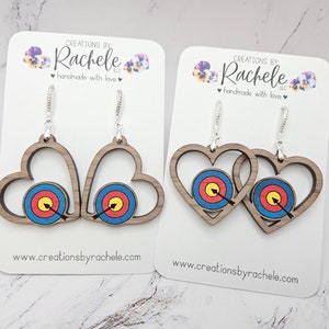 May include: A pair of wooden heart-shaped earrings with a target and arrow design. The earrings are on a white card with the text "CREATIONS BY Rachele LLC handmade with love" and the website "www.creationsbyrachele.com".