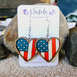 Puede incluir: Un par de pendientes de madera en forma de corazón con un diseño de bandera estadounidense. Los pendientes están en una tarjeta blanca con el texto "Creations by Rachele" y "handmade with love".