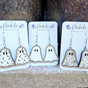 Op de afbeelding: Drie paar Halloween-oorbellen met een spookontwerp. Het eerste paar heeft een wit spook met zwarte vleermuis-silhouetten, het tweede paar heeft een wit spook met zwarte ogen en het derde paar heeft een wit spook met een zwart spinnenweb-ontwerp. Elk paar bevindt zich op een witte kaart met de tekst "CREATIONS BY Rachele LLC handmade with love" en een website-adres.