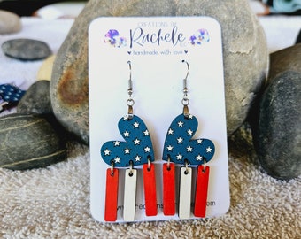 May include: A pair of dangle earrings featuring a red, white, and blue heart with stars and three red, white, and blue stripes hanging below. The earrings are on a white card with the text "CREATIONS BY Rachele LLC handmade with love".