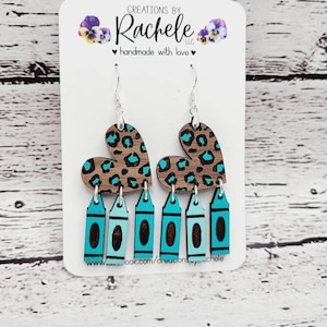 May include: Teal and brown heart-shaped earrings with a leopard print design and teal crayon-shaped charms hanging from the bottom.