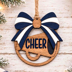May include: A circular wooden ornament featuring a cutout megaphone shape with the word CHEER printed in dark blue. A large decorative bow in navy blue and white sits at the top, attached to a tan jute twine loop for hanging the ornament.