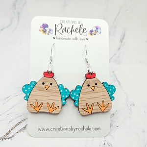 May include: Wooden earrings shaped like a brown chicken with teal wings and white polka dots. The chicken has a red comb and orange feet. The earrings are hanging from silver hooks.
