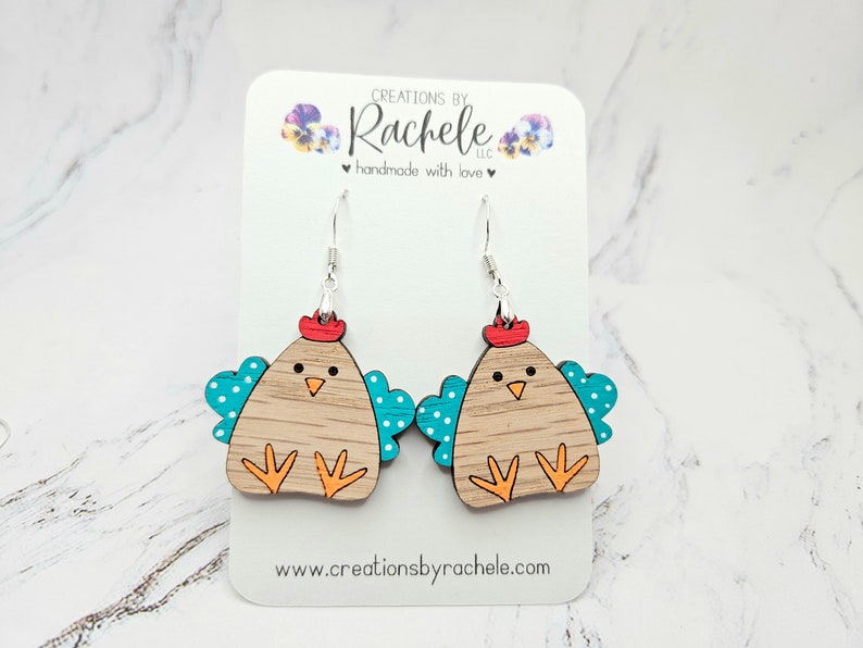 May include: Wooden earrings shaped like a brown chicken with teal wings and white polka dots. The chicken has a red comb and orange feet. The earrings are hanging from silver hooks.