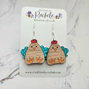 May include: A pair of wooden chicken earrings with teal wings and white polka dots. The earrings are painted with orange feet, a red comb, and brown feathers. The earrings are hanging from silver hooks.