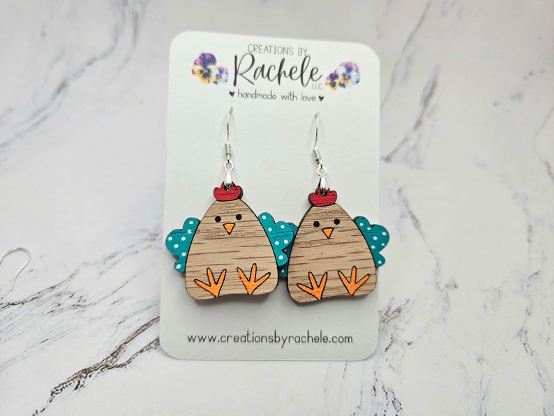 May include: A pair of wooden chicken earrings with teal wings and white polka dots. The earrings are painted with orange feet, a red comb, and brown feathers. The earrings are hanging from silver hooks.