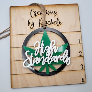 Może przedstawiać: Zielony liść marihuany z napisem "High Standards" białymi literami w czarnym kółku. Ozdoba wisi na drewnianym znaku z napisem "Creations by Rachele".