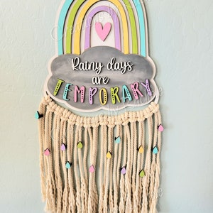 May include: A wooden rainbow wall hanging with a gray cloud that says "Rainy days are TEMPORARY". The rainbow is layered with light blue, green, yellow, purple, and pink. The cloud has a pink heart in the center. The hanging has a fringe of beige yarn with colorful raindrops.