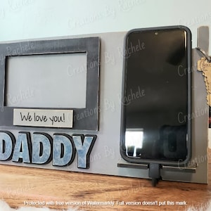 Puede incluir: Un marco de madera gris con un marco de fotos, un soporte para teléfono y un llavero. El marco de fotos tiene un fondo blanco y las palabras "We love you!" escritas en él. El soporte para teléfono es negro y tiene un teléfono en él. El llavero es negro y tiene una llave en él. El marco tiene la palabra "DADDY" escrita en letras azules.