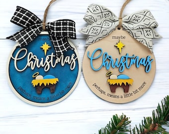 May include: Two round wooden Christmas decorations featuring a blue and white nativity scene with the text "Christmas perhaps, means a little bit more". One decoration has a blue background and the other has a natural wood background. Both decorations have black and white ribbon bows.