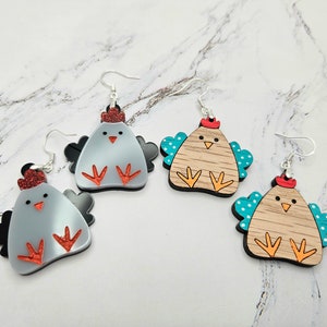 May include: Three pairs of dangle earrings featuring a cartoon chicken design. The chickens are made of wood and painted in various colors, including gray, brown, and turquoise. The earrings have silver hooks.