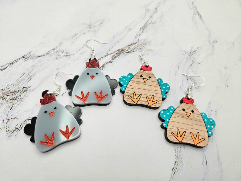 May include: Three pairs of dangle earrings featuring a cartoon chicken design. The chickens are made of wood and painted in various colors, including gray, brown, and turquoise. The earrings have silver hooks.