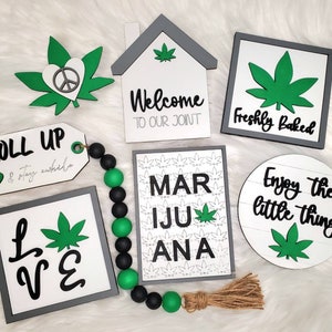 Peut inclure: Une collection de panneaux en bois avec diverses inscriptions sur le thème du cannabis, notamment "Roll Up & Enjoy" et "Welcome to Our Joint". Les panneaux sont décorés de motifs de feuilles de cannabis vertes et sont affichés sur une surface blanche et duveteuse.