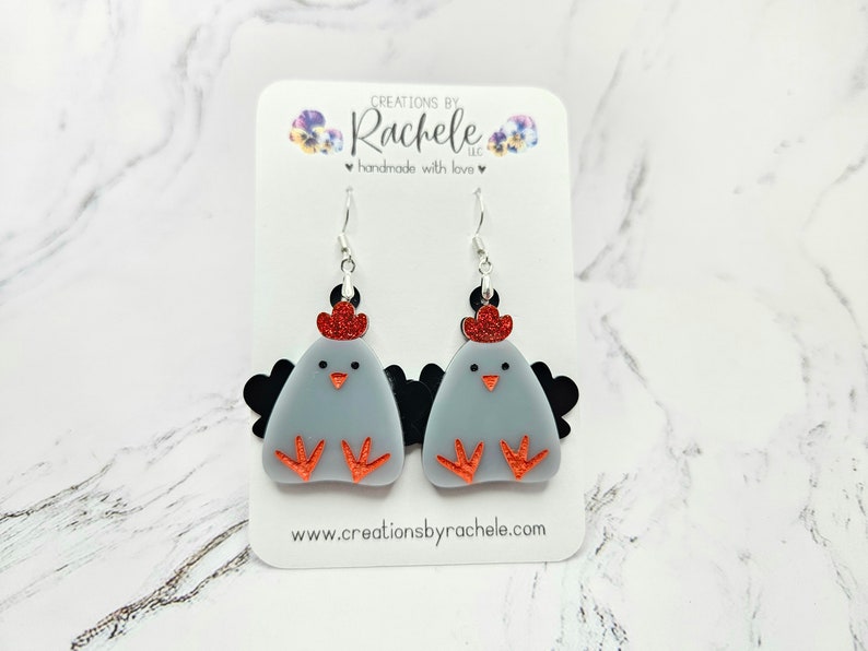 May include: A pair of gray chicken dangle earrings with red glitter accents and black wings. The earrings are on a white card with the text "CREATIONS BY Rachele LLC handmade with love www.creationsbyrachele.com"