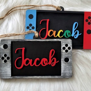 May include: Two wooden ornaments shaped like a Nintendo Switch game console. Both ornaments are black with a gray border and have a red and blue button design. The top ornament has the name "Jacob" written in rainbow colors. The bottom ornament has the name "Jacob" written in red.