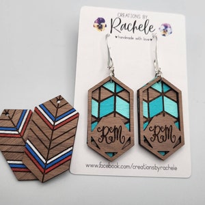 Peut inclure: Une paire de boucles d'oreilles en bois avec un motif géométrique turquoise et marron. Les boucles d'oreilles sont personnalisées avec les initiales "RM" et ont des crochets argentés. Les boucles d'oreilles sont présentées sur une carte blanche avec le texte "Créations by Rachele" et "fait main avec amour".