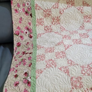 May include: A pink and white quilt with a floral pattern on the border. The quilt is quilted with a swirl pattern.