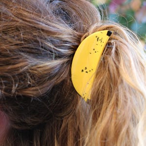 Può includere: Una forcina per capelli a forma di banana gialla con dettagli neri è posta in una coda di cavallo. La forcina ha una punta nera e piccoli segni neri. I capelli sono un mix di marrone e biondo.