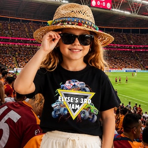 May include: A person wearing a black t-shirt with a monster truck graphic and the text "ELYSE'S 3RD JAM". They are also wearing a straw hat, sunglasses, and white trousers. The background shows a crowded stadium.