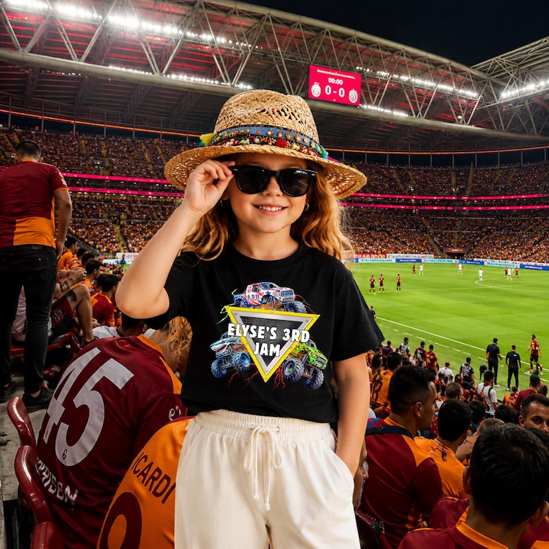 May include: A person wearing a black t-shirt with a monster truck graphic and the text "ELYSE'S 3RD JAM". They are also wearing a straw hat, sunglasses, and white trousers. The background shows a crowded stadium.