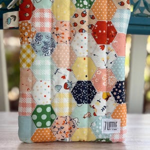 May include: A colorful, quilted pouch with a white zipper and a small fabric tag. The pouch is made of various hexagonal fabric pieces in pastel colors and patterns, including strawberries, florals, and polka dots. The label reads "7 Little".