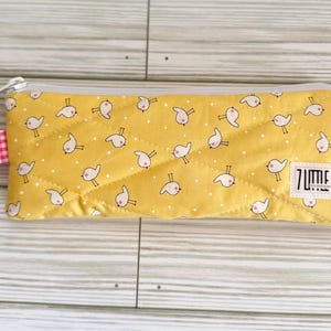 May include: Yellow quilted pencil case with a repeating pattern of white birds with pink accents. The case has a gray zipper, a pink and white gingham pull tab, and a white label that reads "7 LITTLE".