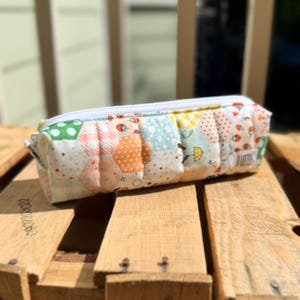 May include: A rectangular pencil case with a white zipper, made of patchwork fabric in various pastel colors. The fabric features hexagonal and other geometric patterns with floral and insect motifs. The case is resting on a wooden pallet.