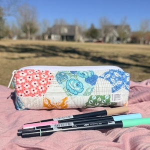 May include: A colorful pencil case with a white zipper, featuring a patchwork design of floral patterns in pink, blue, and orange. Several Tombow ART markers in various colors are arranged in front of the case on a pink cloth.