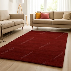 Puede incluir: Una alfombra rectangular de color rojo intenso con una superficie texturizada. La alfombra está colocada sobre un suelo de madera clara en un salón. Dos sofás beige y cojines decorativos son visibles en el fondo.