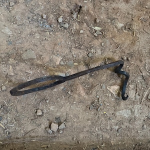 May include: A dark metal branding iron lies on a dirt surface. The iron has a circular handle, a long, textured shaft, and a curved end. The ground is covered in small rocks and debris, with a few scattered pieces of wood.
