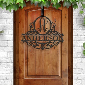 May include: Black metal door sign with the name "ANDERSON" and a decorative "A" initial. The sign is suspended from a black hook and hangs on a wooden door. The design includes ornate scrollwork.