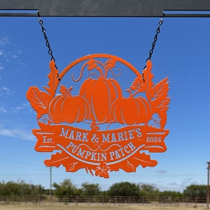 May include: Orange metal sign with a pumpkin patch theme, hanging from a black chain. The sign features three pumpkins and the text "Mark & Marie's Pumpkin Patch Est. 2024" in a decorative font. The background is a blue sky.