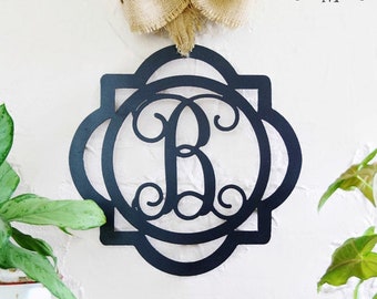 May include: Black metal monogram with a decorative scroll design and a single initial "B" in the center. The monogram is hanging on a white wall with a burlap bow above it.