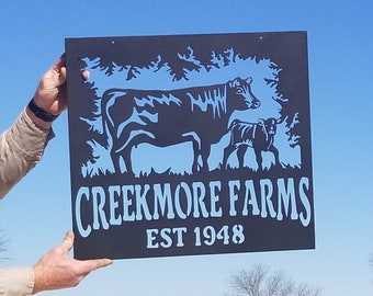 May include: A black square sign with a light blue cow and calf silhouette design, with a tree outline in the background. The sign reads "CREEKMORE FARMS EST 1948" in white lettering. The sign is held up against a blue sky.