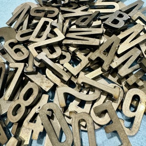 May include: A large pile of metal letters and numbers in a variety of styles. The letters and numbers are all in a gold color and have a slightly distressed finish.