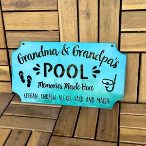 May include: A teal blue metal sign with the text "Grandma & Grandpa's POOL Memories Made Here KEEGAN, ANDREW, ALEXIS, JACK AND MAISIE" in black lettering. The sign has a decorative border and cutouts of flip flops and a snorkel.