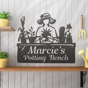 May include: Black metal silhouette wall art featuring a woman gardening with plants and the text "Marcie's Potting Bench".