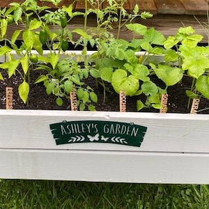 May include: A white wooden raised garden bed with a green sign that says "Ashley's Garden". The bed is filled with soil and various plants, including tomatoes, peppers, zucchini, squash, and herbs. Each plant has a wooden stake with a handwritten label.