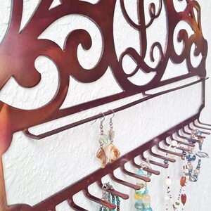 K&ouml;nnte beinhalten: Ein kupferfarbener Schmuckorganizer aus Metall mit einem dekorativen Schn&ouml;rkeldesign und einem einzigen Initial "L" in der Mitte. Der Organizer hat ein Regal und mehrere Haken zum Aufh&auml;ngen von Halsketten, Armb&auml;ndern und Ohrringen.