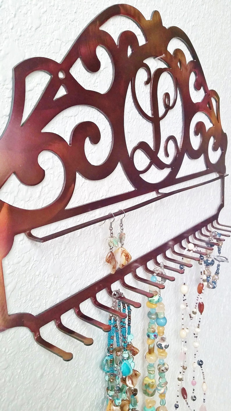 K&ouml;nnte beinhalten: Ein kupferfarbener Schmuckorganizer aus Metall mit einem dekorativen Schn&ouml;rkeldesign und einem einzigen Initial "L" in der Mitte. Der Organizer hat ein Regal und mehrere Haken zum Aufh&auml;ngen von Halsketten, Armb&auml;ndern und Ohrringen.